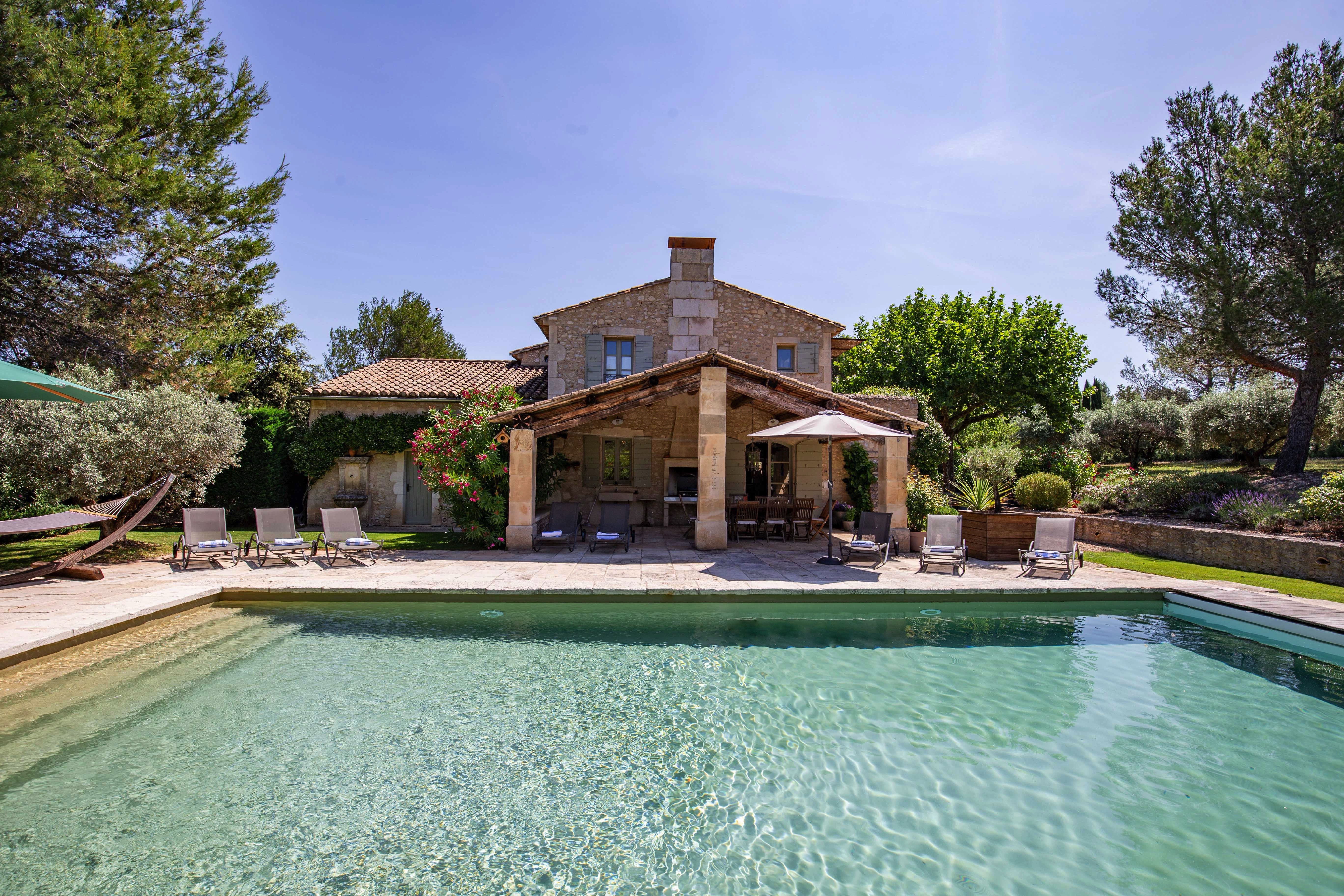 A luxurious stone villa with a large pool in the foreground, surrounded by lush greenery, under a clear blue sky. Loungers and a shaded patio area offer relaxation spots.